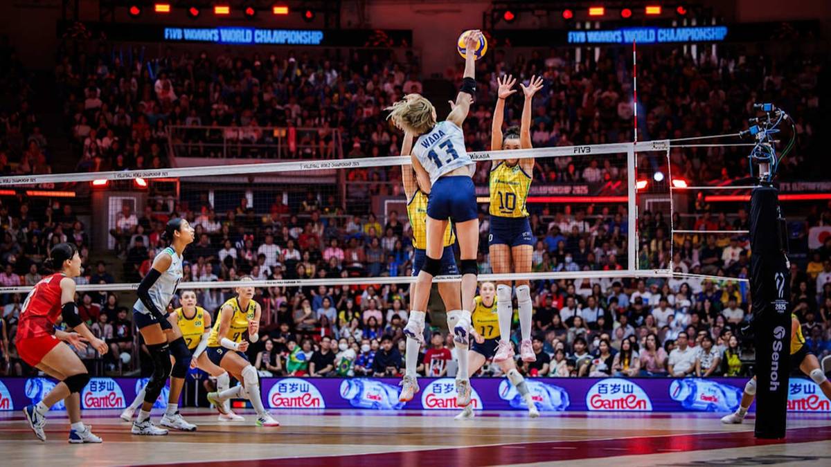 Zawodniczki siatkówki grają mecz na tle publiczności na trybunach. Jedna z zawodniczek w białej koszulce z numerem 13 wyskoczyła do zagrania piłki nad siatką, podczas gdy zawodniczka w żółtej koszulce z numerem 10 również próbuje ją zablokować. W tle widać kolejne zawodniczki i dużą widownię.