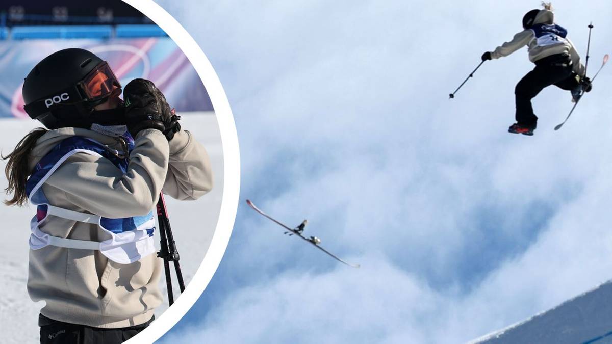 Dziewczynka w kasku i goglach narciarskich, trzymająca aparat fotograficzny na statywie, patrzy w kierunku skoczni narciarskiej. W tle narciarz wykonuje skok.