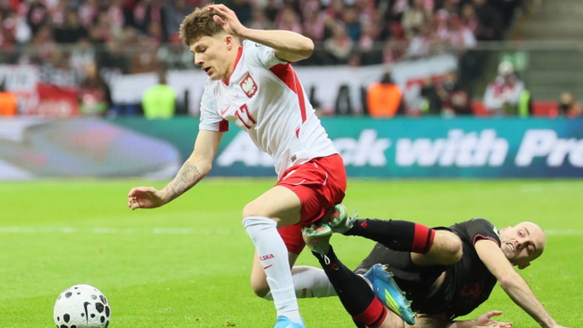 Piłkarz w białej koszulce walczy o piłkę z zawodnikiem leżącym na murawie.