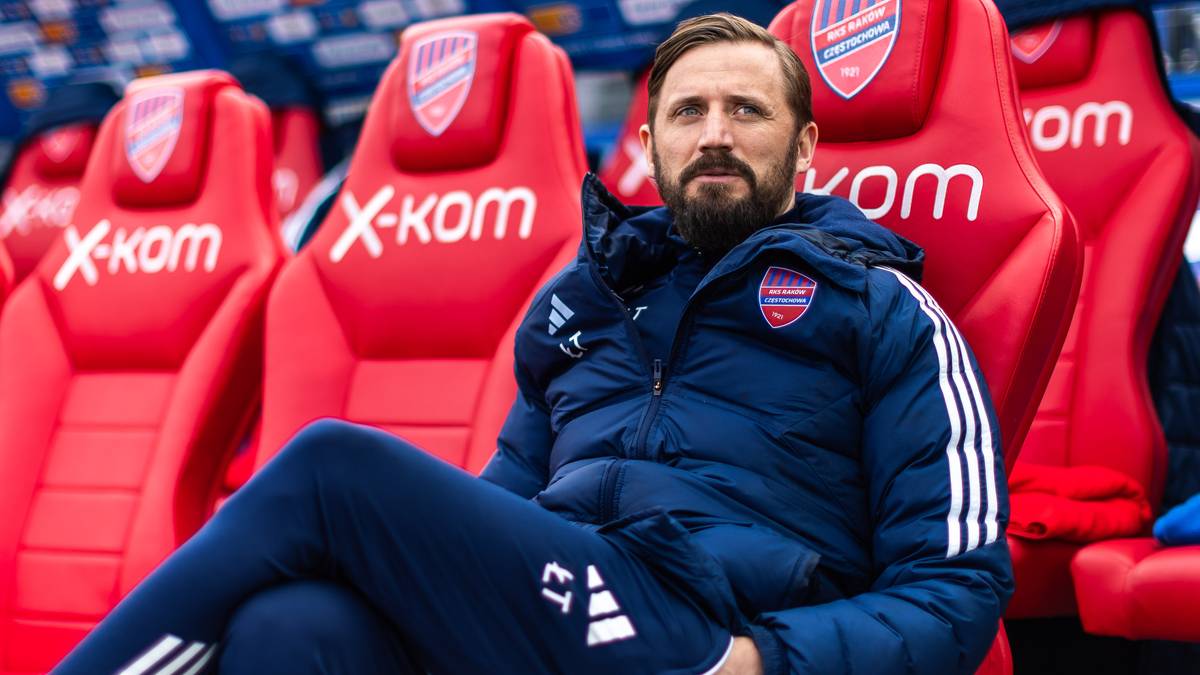 Mężczyzna w stroju sportowym siedzi na czerwonym krześle na tle stadionu.