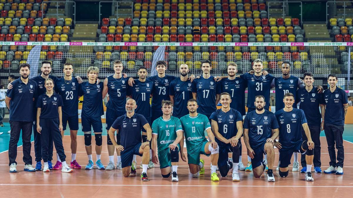 Drużyna siatkarska w niebieskich i zielonych strojach sportowych pozuje do zdjęcia na tle trybun hali sportowej. Zawodnicy stoją i kucają w dwóch rzędach.