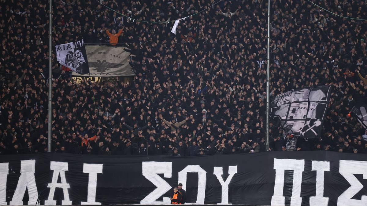 Tłum kibiców na trybunach stadionu, widoczne flagi i transparenty z napisami.