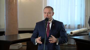 Mężczyzna w ciemnym garniturze i bordowym krawacie przemawia do mikrofonu, gestykulując obiema rękami. W tle widać meble i okno.