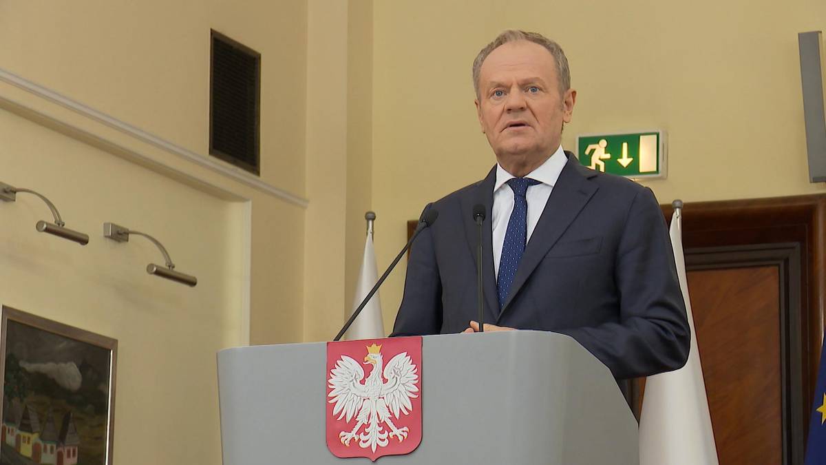 Premier Donald Tusk przemawia przy m&oacute;wnicy z godłem Polski.