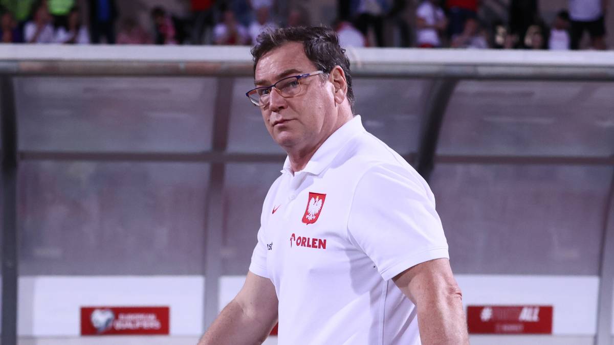 Mężczyzna w białej koszulce polo z herbem Polski, w okularach, na tle stadionu.