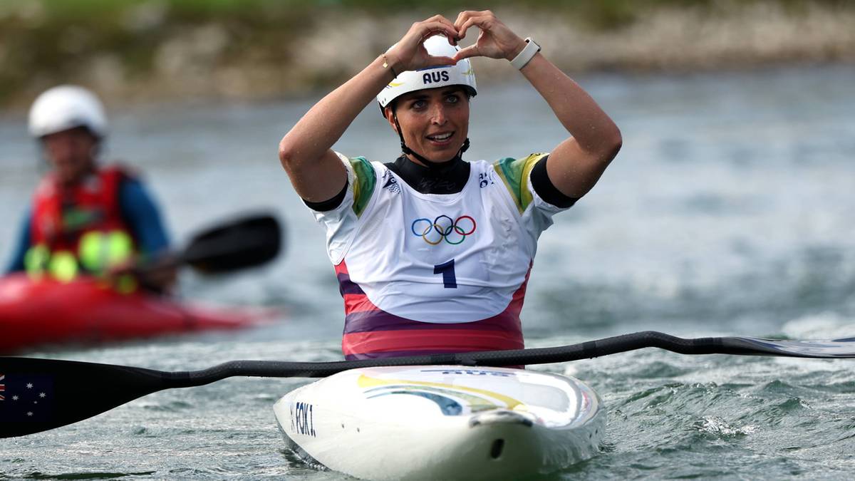 Kobieta w kajaku, w kasku i stroju sportowym z numerem "1" na piersi, tworzy serce z dłoni nad głową.