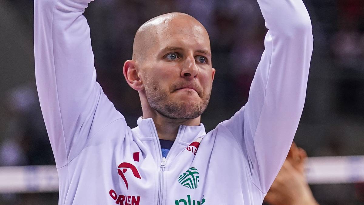 Portret siatkarza Bartosza Kurka z uniesionymi rękami, ubranego w białą bluzę z logo.