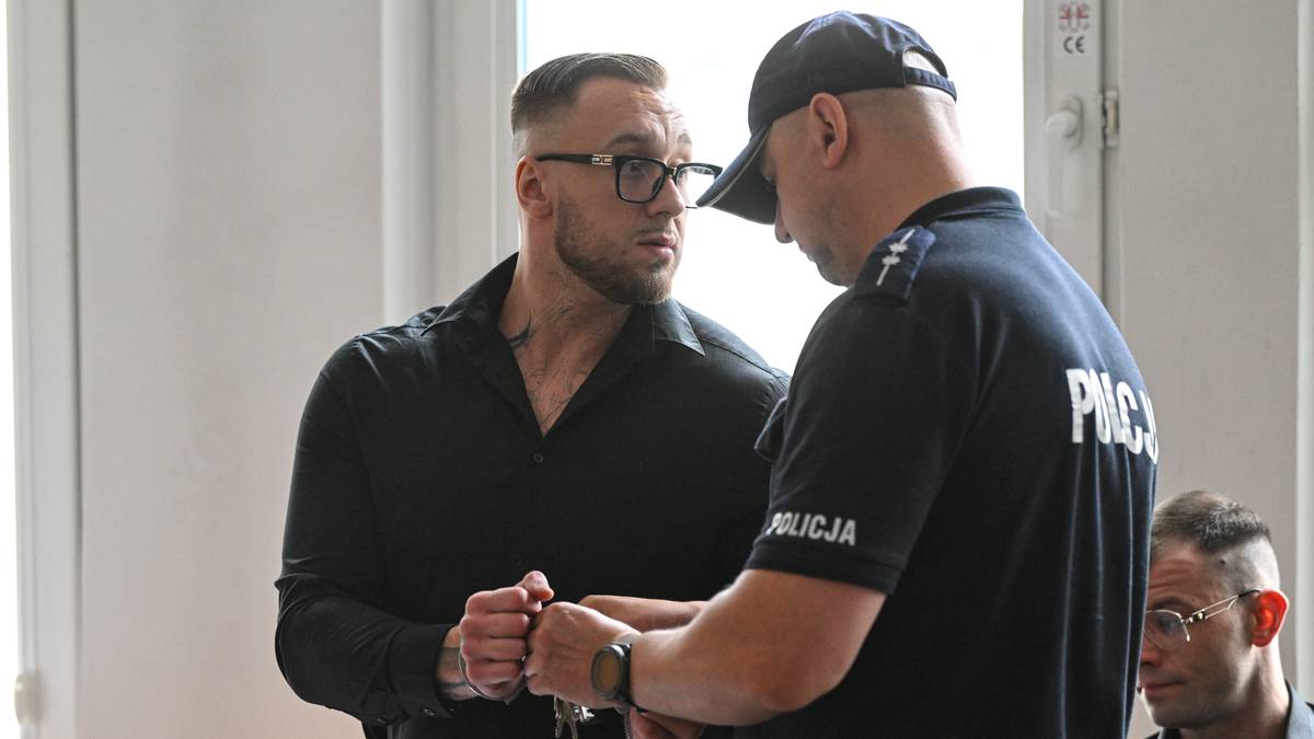 Mężczyzna w czarnej koszuli i okularach jest prowadzony przez policjanta w mundurze.