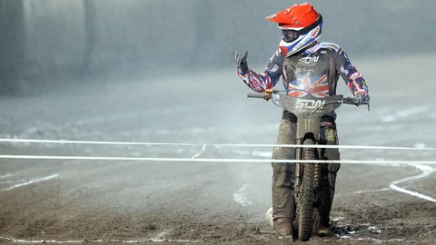 Żużlowiec w czerwono-białym kasku i kombinezonie z brytyjską flagą, stojący na motocyklu na błotnistym torze żużlowym, unosi rękę w geście pozdrowienia.
