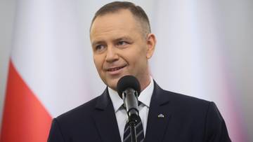 Mężczyzna w garniturze przemawia do mikrofonu, na tle flagi Polski.