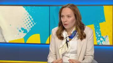 Kobieta w jasnej marynarce i białej koszuli, ubrana w apaszkę z niebieskimi i ż&oacute;łtymi elementami, siedzi i m&oacute;wi. Tło stanowią dwa ekrany z abstrakcyjnymi, geometrycznymi wzorami w kolorach niebieskim i ż&oacute;łtym.