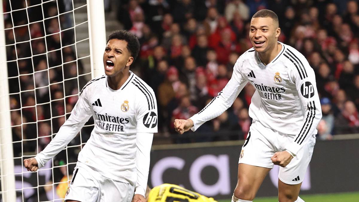 Dw&oacute;ch piłkarzy w białych strojach świętuje zdobycie bramki, bramkarz leży na ziemi, piłka w siatce, kibice na trybunach.