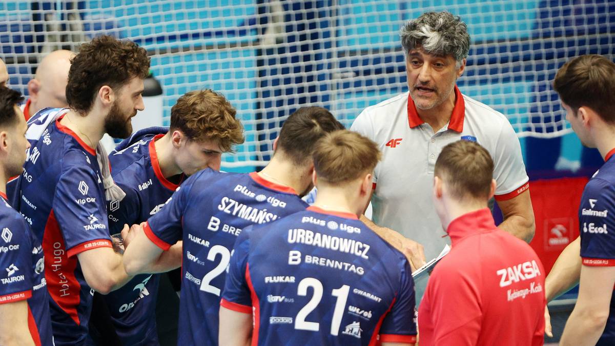 Trener siatk&oacute;wki w białej koszulce m&oacute;wi do grupy zawodnik&oacute;w w niebieskich strojach, kt&oacute;rzy go otaczają i słuchają z uwagą.