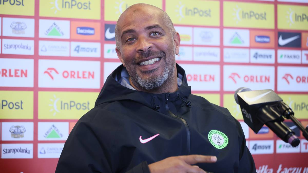 Trener reprezentacji Nigerii z brodą, ubrany w czarną bluzę z kapturem z logo Nike i okrągłym emblematem na piersi, uśmiecha się do mikrofonu podczas konferencji prasowej.