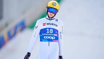 Skoczek narciarski w białym kombinezonie, żółtym kasku i niebieskich goglach stoi na śniegu.