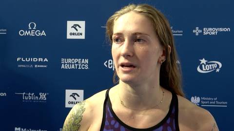 Portret Dominiki Sztandery, polskiej pływaczki, podczas wywiadu. Kobieta ma jasne włosy, spięte w luźny kucyk, i lekko zmęczony wyraz twarzy. Jest ubrana w str&oacute;j kąpielowy. W tle widoczne są logotypy sponsor&oacute;w i organizator&oacute;w zawod&oacute;w.