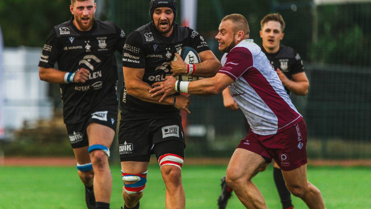 Rugbiści w akcji, jeden zawodnik w ciemnym stroju z piłką jest atakowany przez rywala w jasnym stroju.