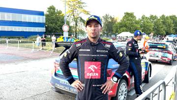 Szymon Gospodarczyk stoi przed samochodem rajdowym, ubrany w kombinezon i czapkę.