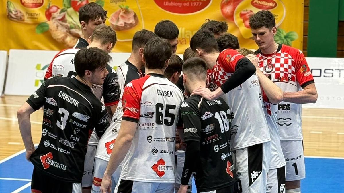 Drużyna siatkarzy w białych i czarnych strojach sportowych stoi w k&oacute;łku na boisku halowym.