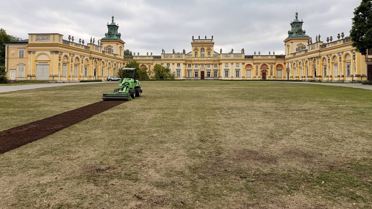 Widok na Pałac Kr&oacute;la Jana III w Wilanowie z dziedzińca przed budynkiem. Widać zielony trawnik, na kt&oacute;rym pracuje zielona kosiarka lub glebogryzarka, tworząc ciemną ścieżkę. W tle znajduje się okazała, ż&oacute;łta fasada pałacu z zielonymi dachami i wieżyczkami.