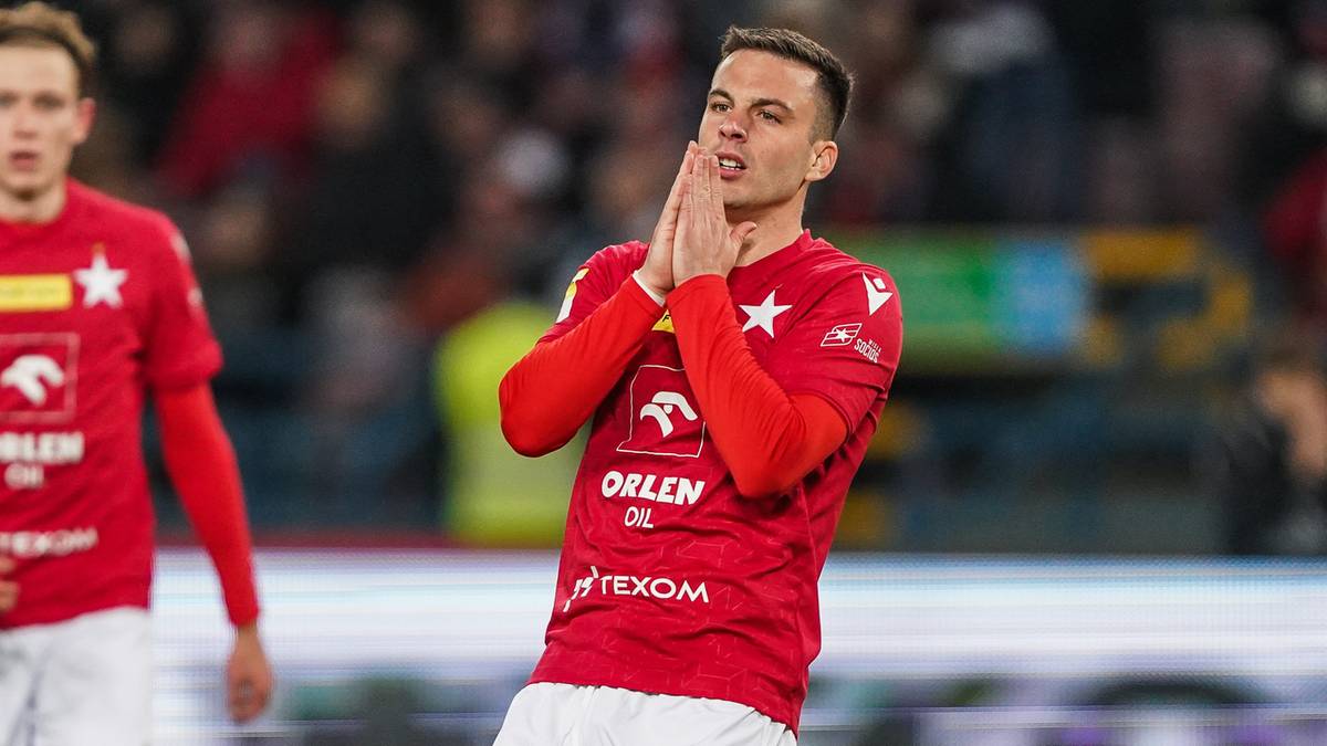 Piłkarz w czerwonej koszulce z białym herbem klubu i napisem "ORLEN OIL" na piersi, klęczy z rękami złożonymi przed twarzą, wyrażając emocje na tle trybun.