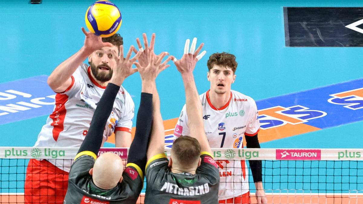 Siatkarze w akcji podczas meczu, jeden z nich serwuje piłkę, podczas gdy pozostali pr&oacute;bują ją zablokować.