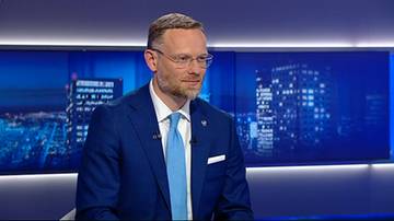 Mężczyzna w garniturze i w okularach wypowiada się w studio telewizyjne na tle niebieskich ekran&oacute;w.