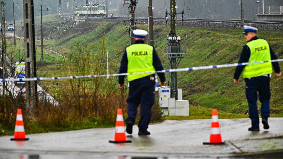 Dwóch policjantów w kamizelkach odblaskowych zabezpiecza teren wokół uszkodzonych torów kolejowych. W tle widać pociąg i infrastrukturę kolejową.