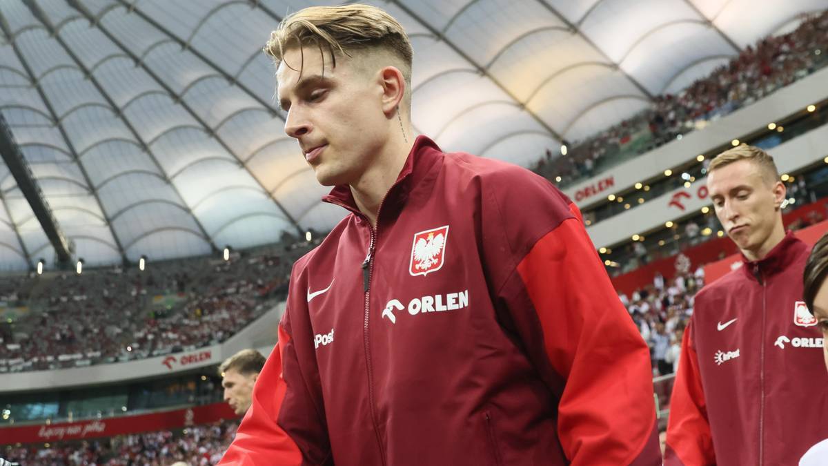 Piłkarz reprezentacji Polski w bordowej bluzie z czerwonymi rękawami i białym logo wchodzi na boisko stadionu. W tle widać trybuny wypełnione kibicami.