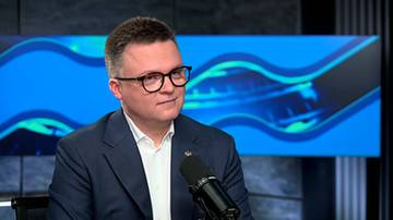 Szymon Hołownia w marynarce i okularach siedzi przed mikrofonem na tle niebieskiego ekranu.