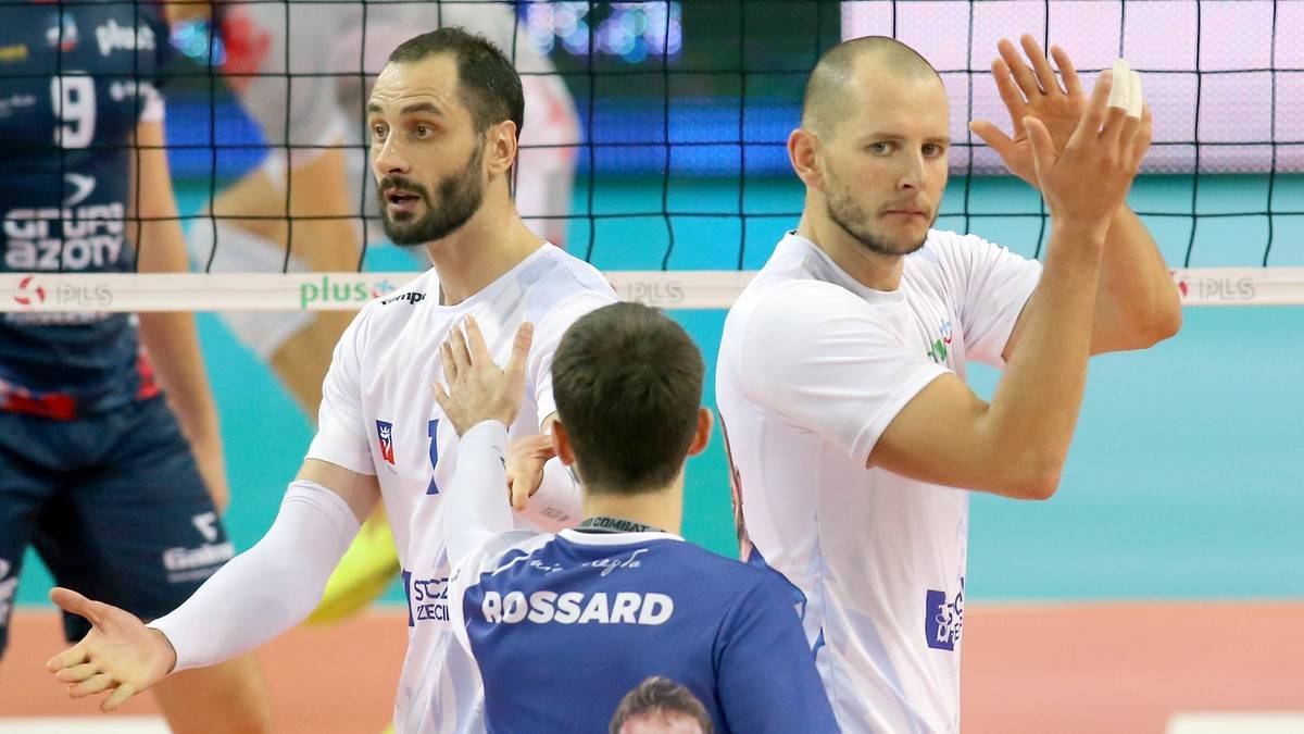 Dwóch siatkarzy w białych koszulkach stoi obok siebie i rozmawia. W tle widać siatkę i innych zawodników.