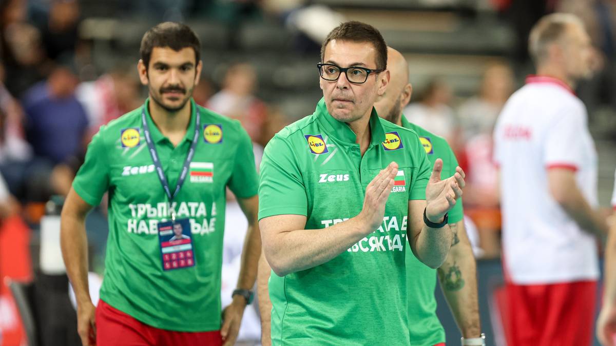 Dwóch trenerów siatkówki w zielonych koszulkach z identyfikatorami na szyi stoi obok siebie. Jeden z nich, bliżej przodu, w okularach, klaszcze, patrząc w bok. Drugi, z brodą, stoi z lewej strony, z opuszczonymi rękami.