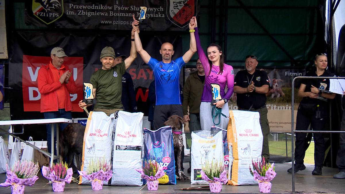 Na podium stoją zwycięzcy konkursu ps&oacute;w. Mężczyzna w niebieskiej koszulce w środku trzyma dwa puchary. Obok niego stoją inne osoby z nagrodami. Na pierwszym planie widać worki z karmą dla ps&oacute;w i kwiaty.