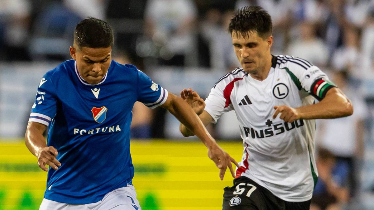 Dwóch piłkarzy na boisku, jeden w niebieskiej koszulce z napisem "FORTUNA", drugi w białej koszulce z napisem "PLUS500".