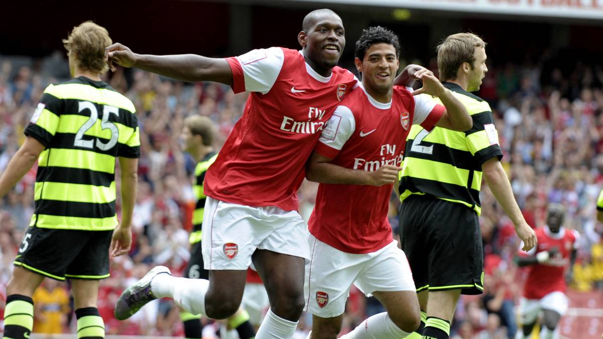 Dwóch piłkarzy w czerwonych strojach Arsenalu ściska sobie ręce i uśmiecha się do siebie, podczas gdy inni piłkarze w czarno-żółtych strojach stoją w tle.