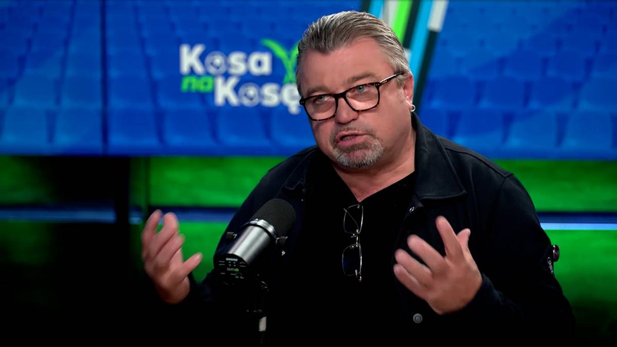 Mężczyzna w okularach, z siwymi włosami, w czarnej koszuli, gestykuluje dłońmi podczas wypowiedzi do mikrofonu.