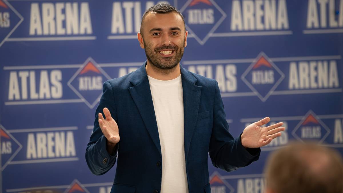 Mężczyzna w granatowej marynarce i białej koszulce, z uniesionymi dłońmi, przemawia na tle niebieskiej ściany z logotypami.