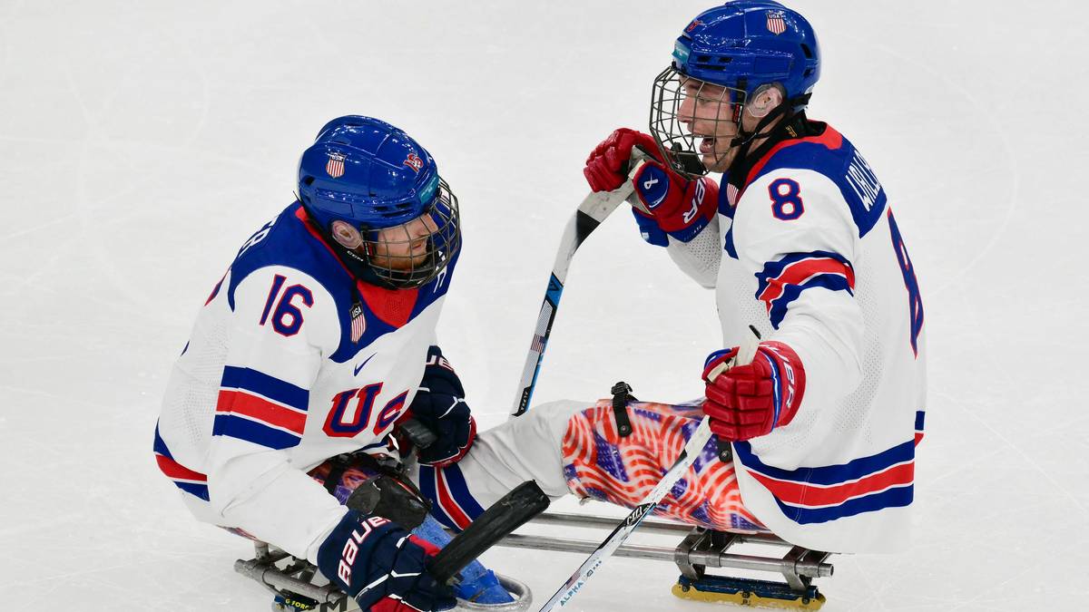 Dw&oacute;ch zawodnik&oacute;w w niebieskich kaskach i białych strojach drużyny USA z numerami 16 i 8 siedzi na sankach hokejowych i trzyma kije.