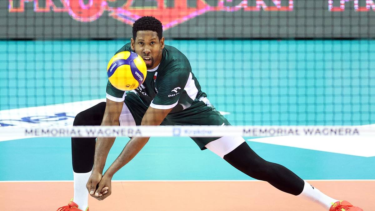 Siatkarz w zielonej koszulce z ż&oacute;łtymi wstawkami pr&oacute;buje przyjąć piłkę w biegu na boisku.