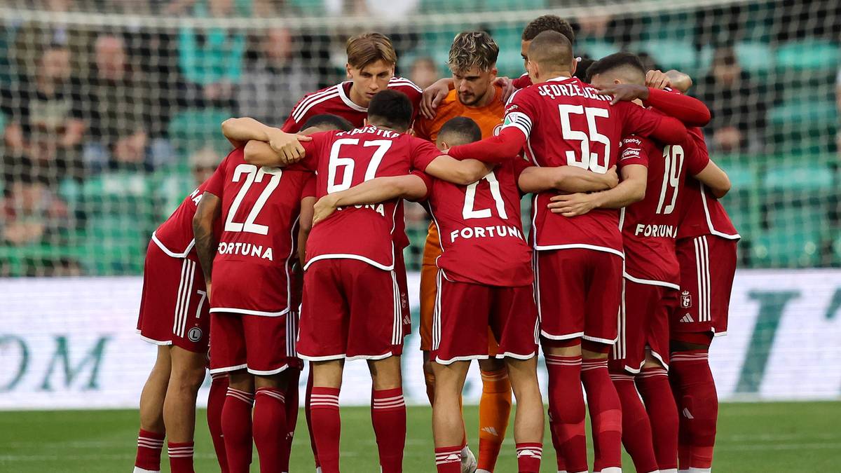 Grupa piłkarzy w czerwonych strojach stoi w kręgu, przytulając się i rozmawiając.