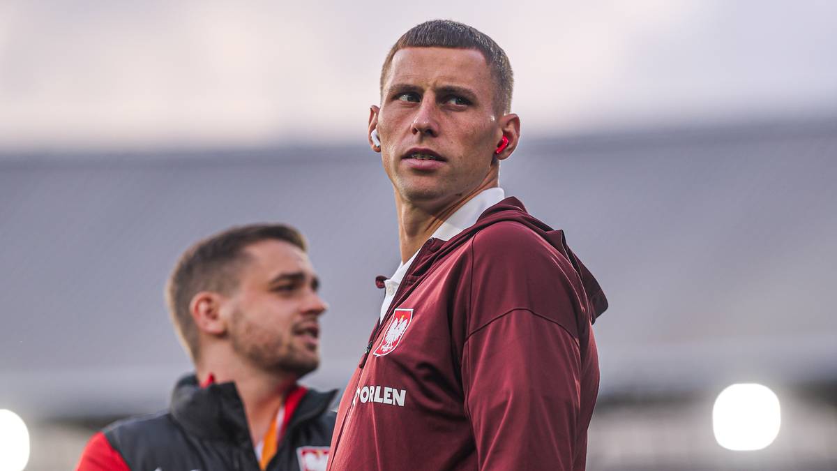 Młody mężczyzna o krótkich włosach, ubrany w bordową bluzę z białym godłem, stoi na zewnątrz. Z tyłu widać drugiego mężczyznę w ciemnej kamizelce. Niebo jest zachmurzone.