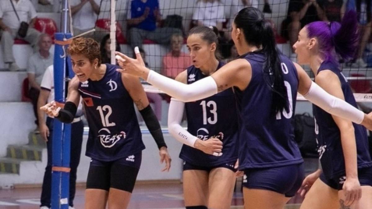 Cztery siatkarki w granatowych strojach podczas meczu siatkówki, jedna z nich z numerem 12 wykonuje gest ręką.