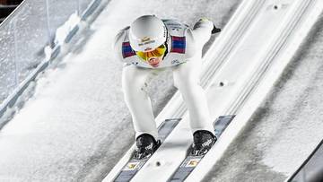 Skoczek narciarski w białym stroju z goglami na nosie zjeżdża po rozbiegu skoczni narciarskiej.