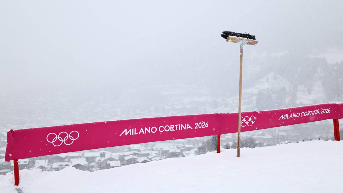 Banda sponsorska "MILANO CORTINA 2026" na ośnieżonym stoku z kółkami olimpijskimi, obok miotła na wysięgniku.