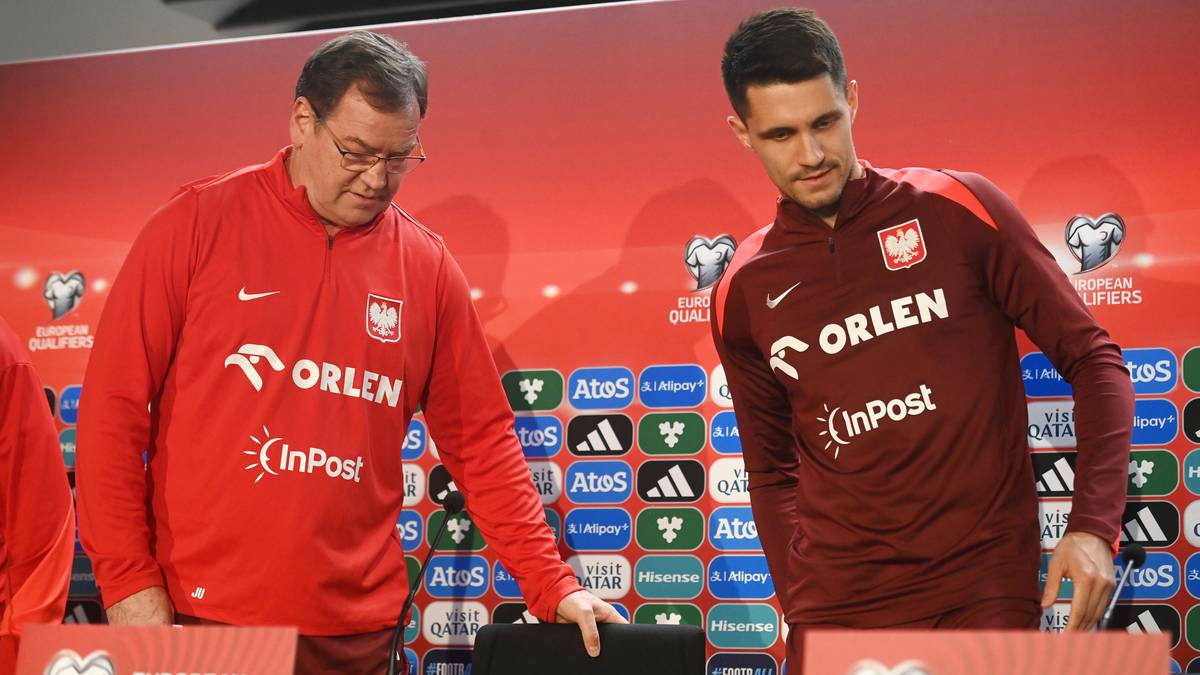 Dwóch mężczyzn w strojach reprezentacji Polski, jeden starszy w czerwonej bluzie, drugi młodszy w bordowej koszulce, stoją przed ścianką z logotypami.