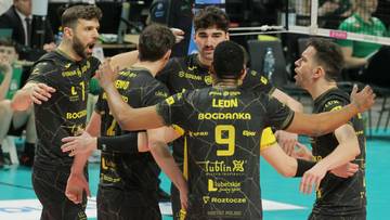 Grupa siatkarzy w czarnych strojach z ż&oacute;łtymi detalami świętuje na boisku, tworząc zwartą grupę.