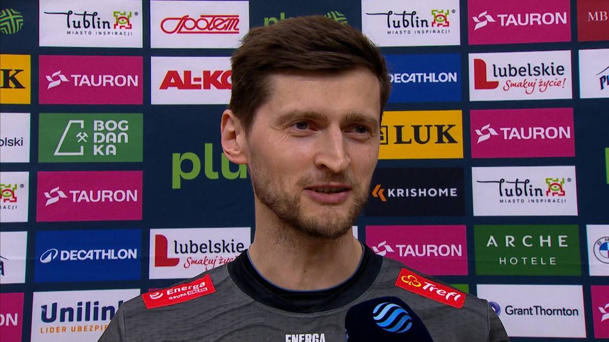 Portret Piotra Nowakowskiego, mężczyzny z zarostem i krótkimi włosami, na tle kolorowej ścianki sponsorskiej.