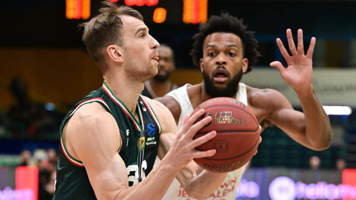 Dw&oacute;ch koszykarzy walczy o piłkę podczas meczu. Zawodnik w zielonym stroju prowadzi piłkę, a zawodnik w białym stroju pr&oacute;buje go zatrzymać.