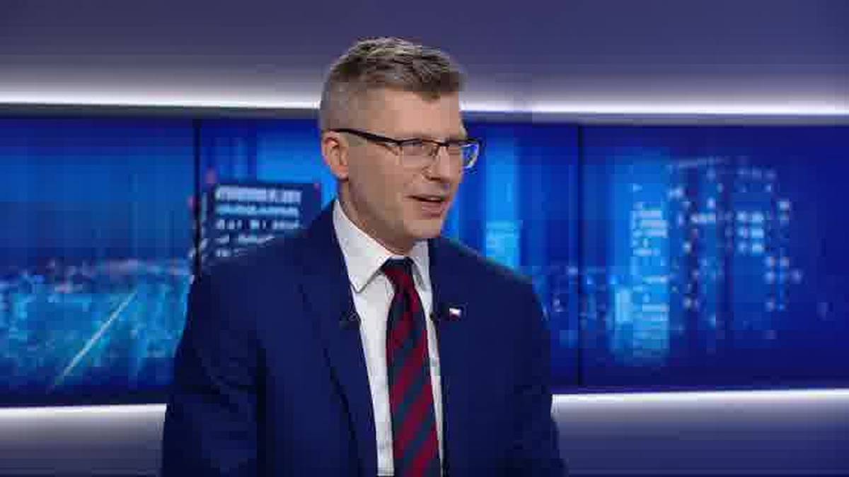 Mężczyzna w garniturze i krawacie m&oacute;wi do kamery, w tle niebieskie ekrany z grafikami miasta.