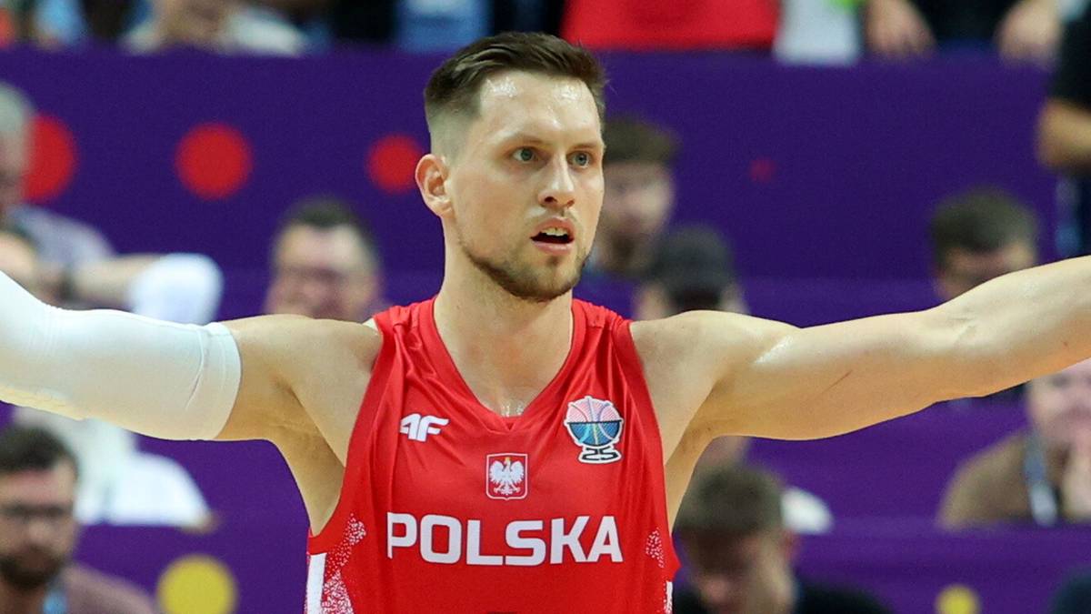 Portret koszykarza w czerwonej koszulce z napisem POLSKA. Zawodnik patrzy w bok, jego prawa ręka jest lekko uniesiona.